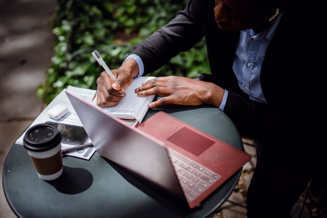 A human writes on a pad of paper, next to a laptop