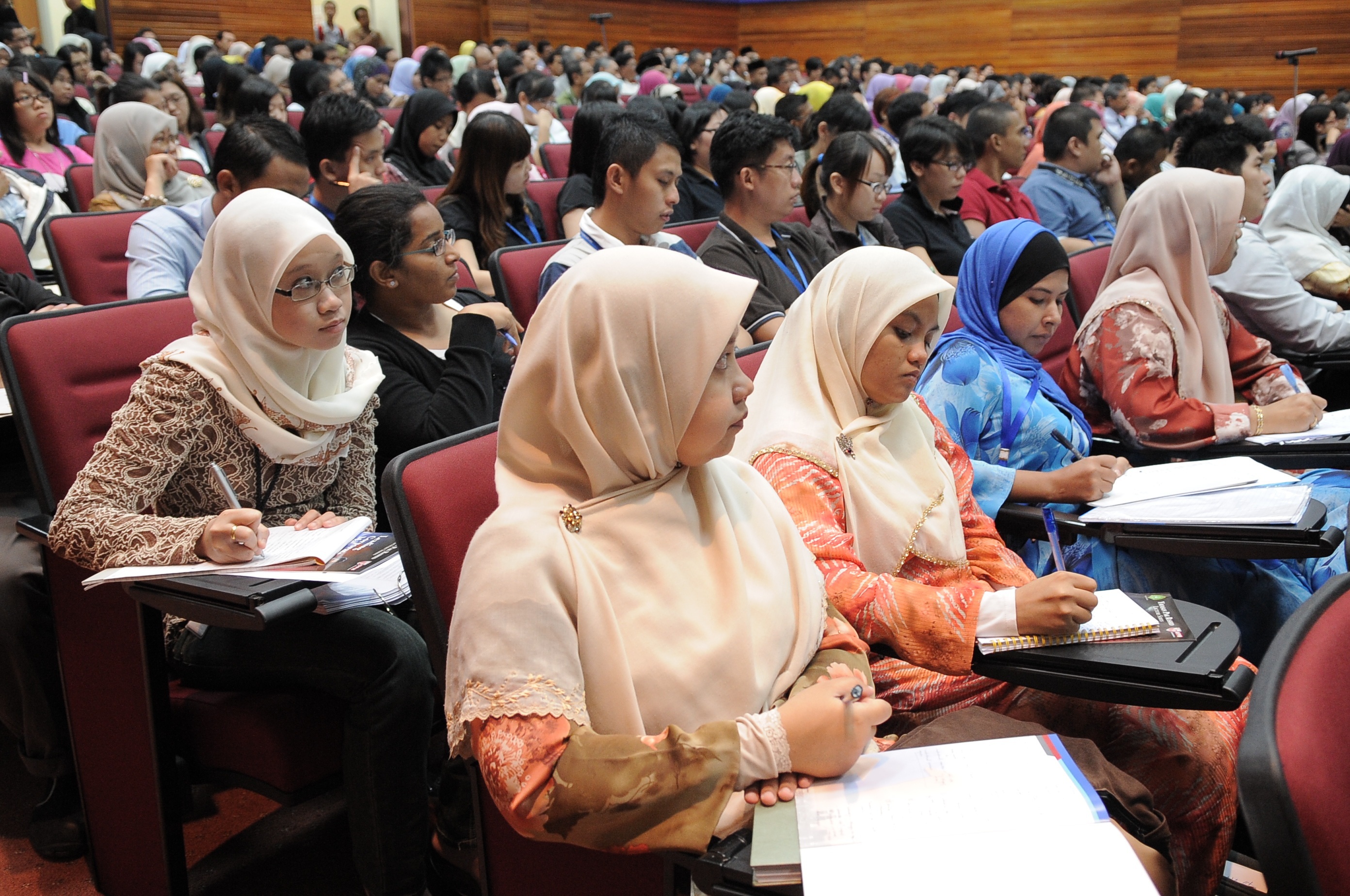 A large university lecture hall filled with students taking notes; the foreground of the image features many young women dressed in hijab.