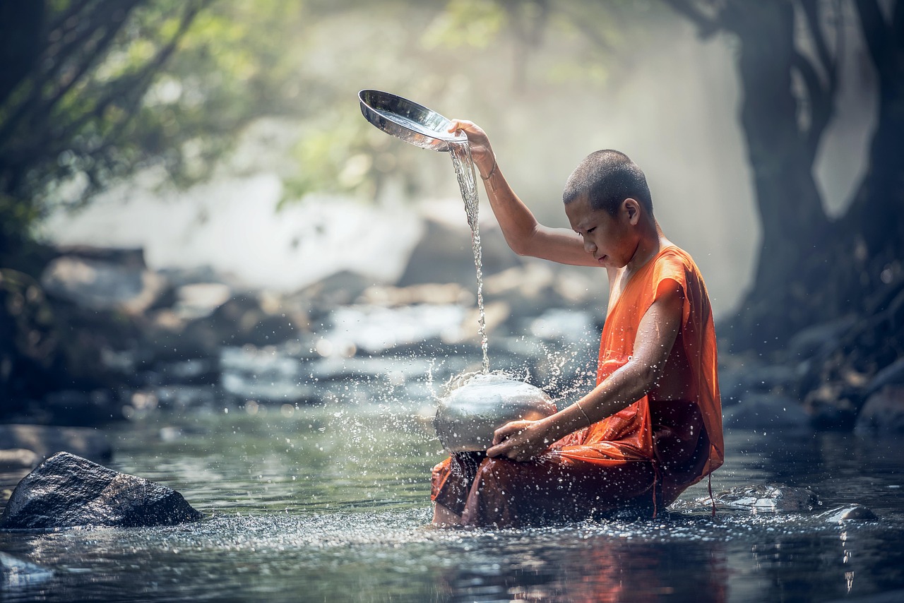 A Tibetan boy in orange robes sits in a stream spilling water from a shallow dish