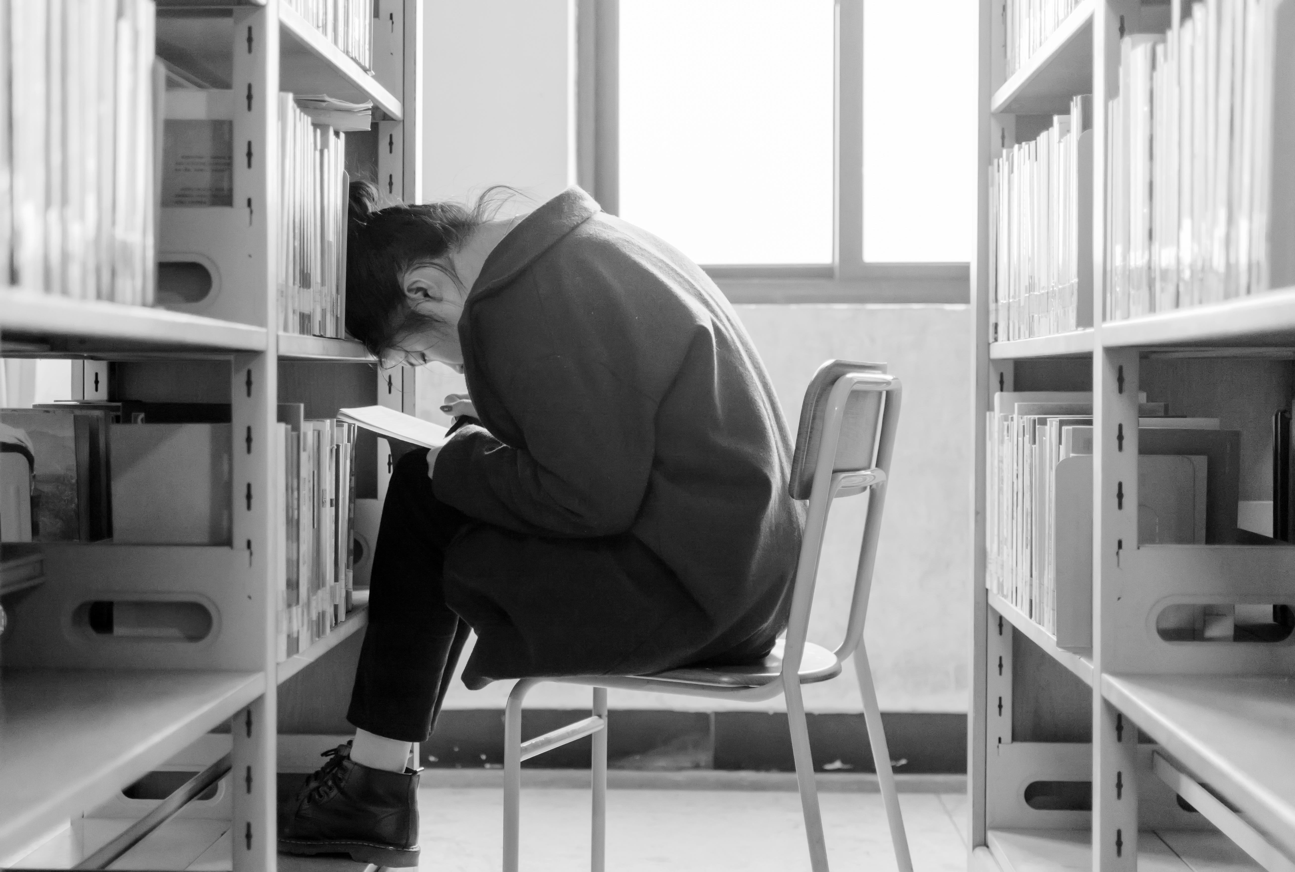 A black-and-white image of a young woman sitting while crouching over a notebook surrounded by stacks of library books