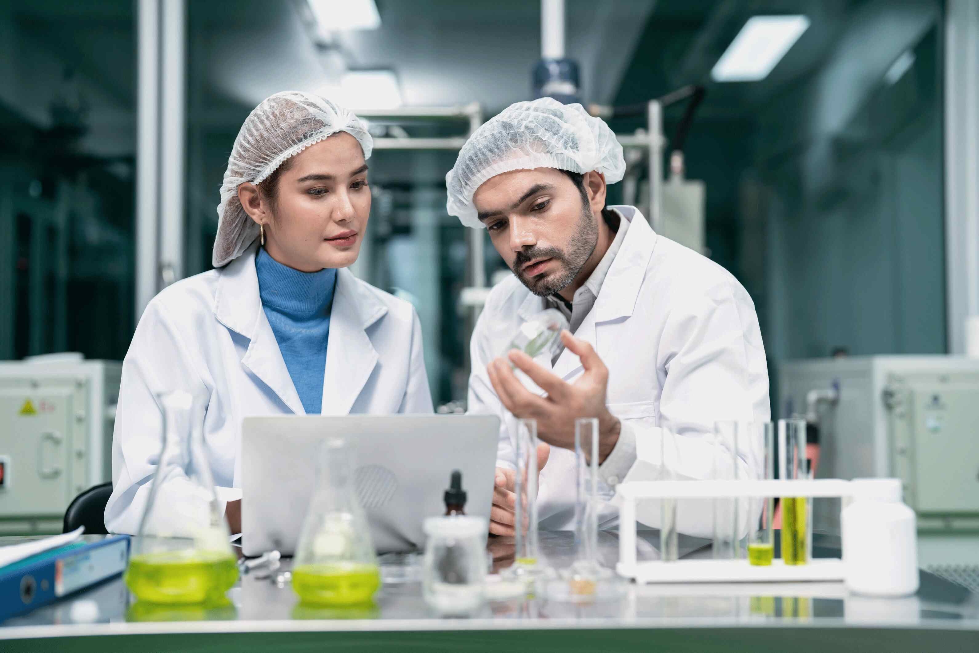 A young man and woman in a laboratory wearing white coats and hair caps looking at a petri dish