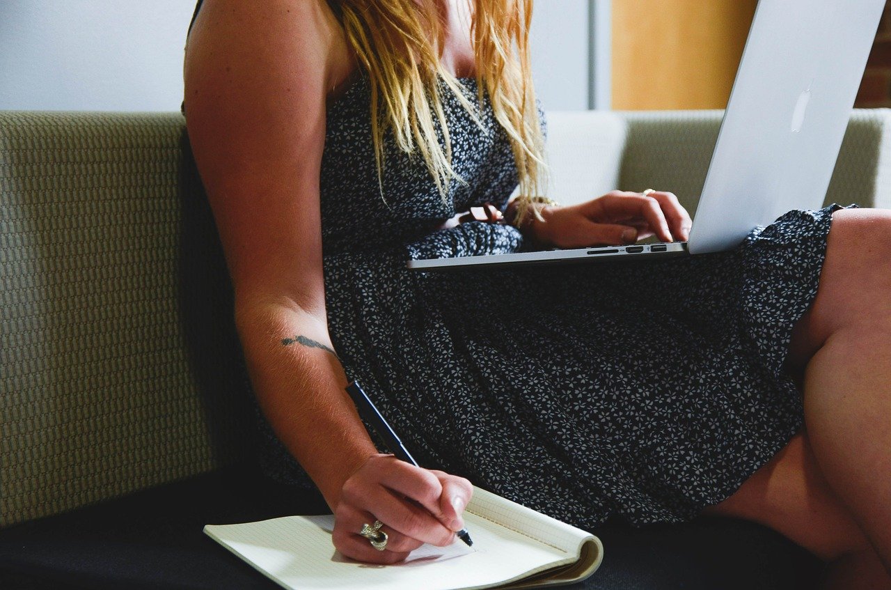 A woman balances a laptop on her lap, while taking notes in a pad.